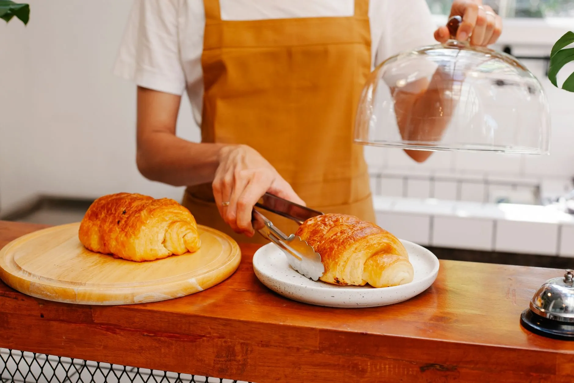 Hands in an orange apron lifting a croissant with tongs at a bakery counter
