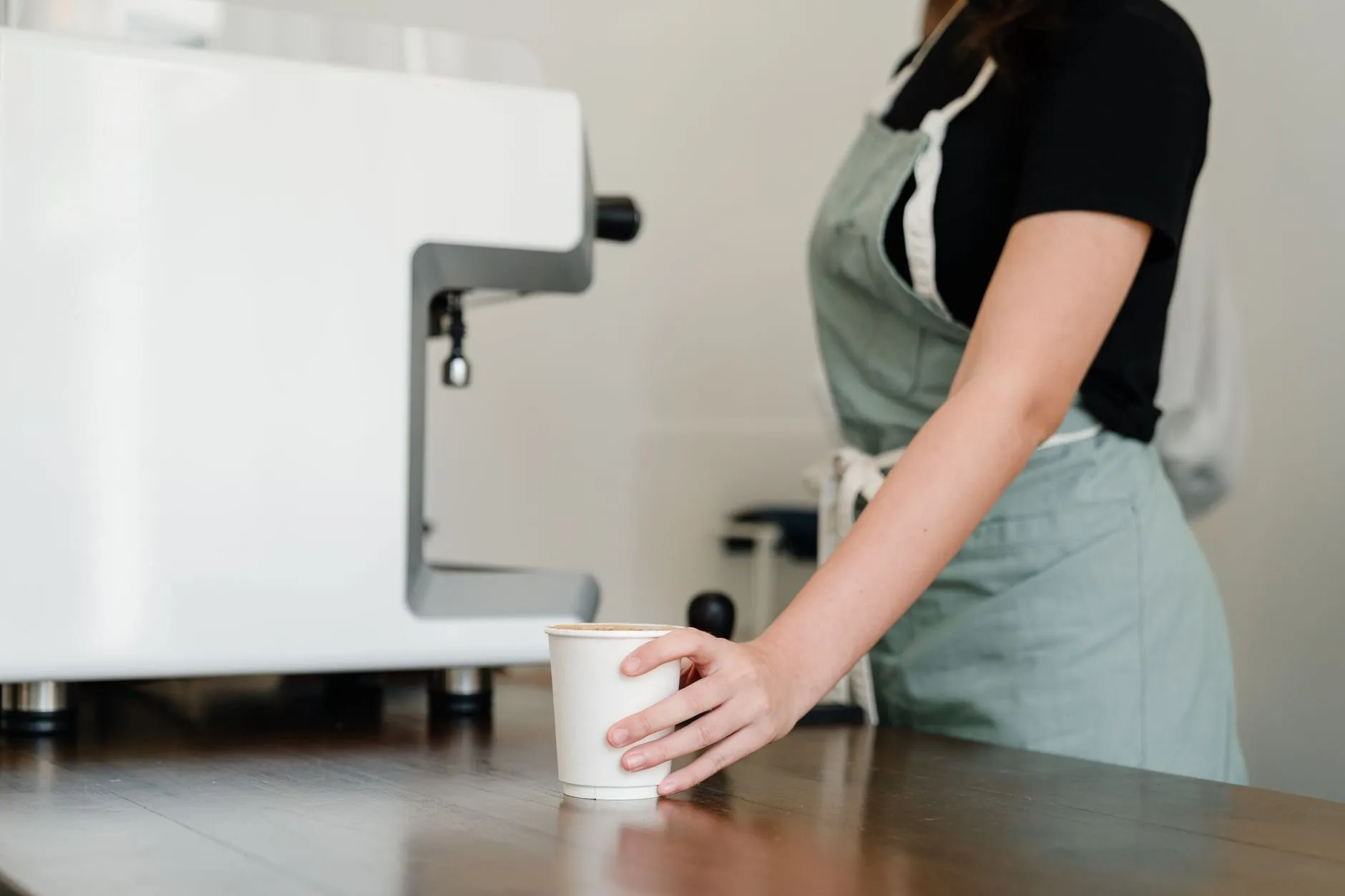 A barista in an apron sets a takeaway cup on the counter next to the espresso machine