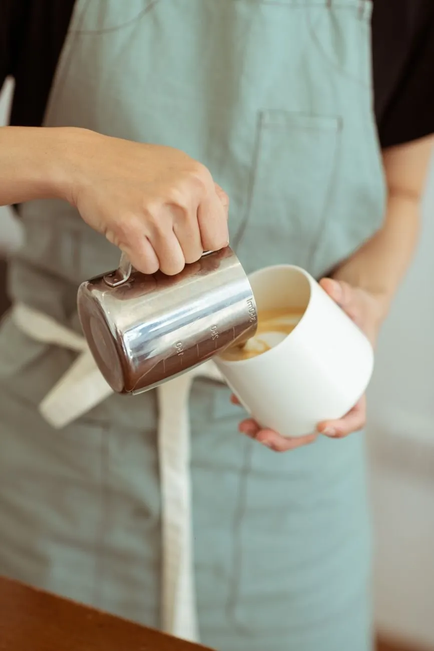 A barista in a sage apron pouring steamed milk into a cup