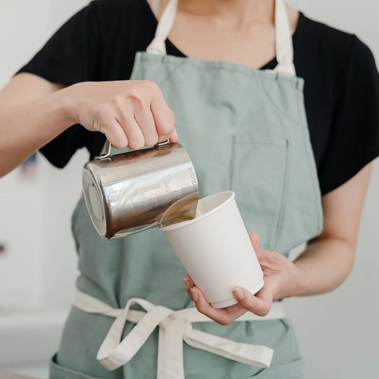 A barista in a sage apron pouring steamed milk into a paper cup